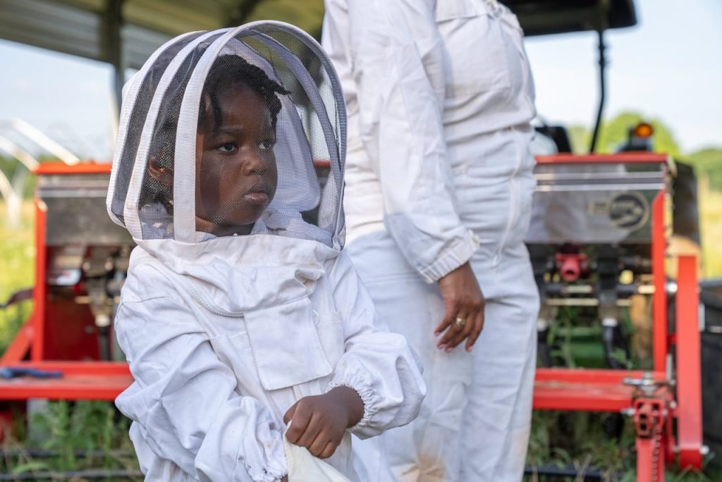 A young child in protective beekeeping clothing stands outdoors during a sunny day.